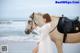 A woman in a white dress standing next to a horse on the beach.