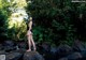 A woman in a black bathing suit standing on a rock by a stream.